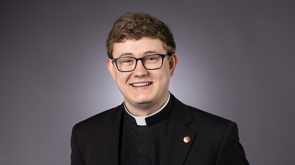 Portrait of a smiling male faculty member wearing a clerical collar and glasses against a gray background.